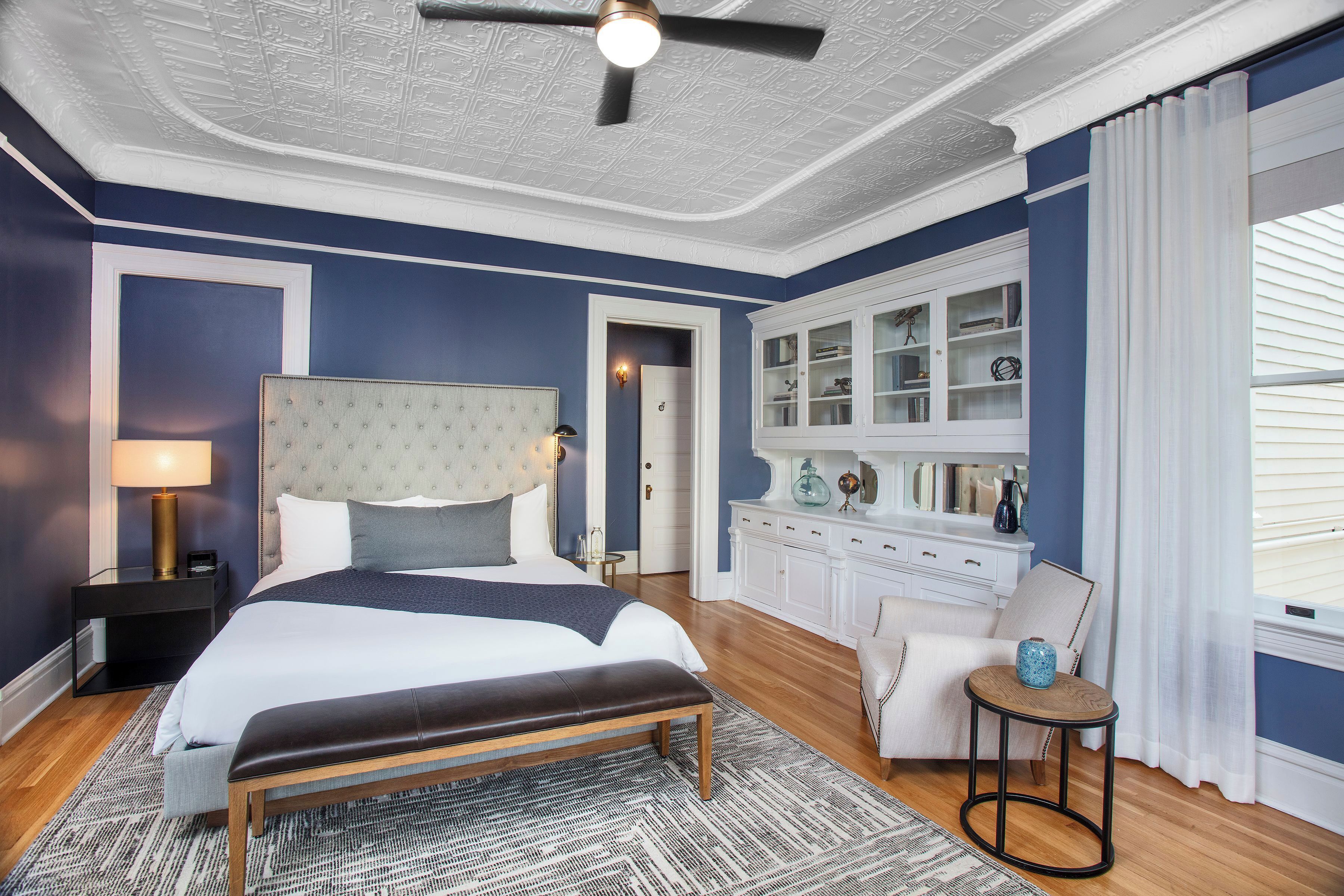 A bedroom with a large bed, a bedside table with a lamp, a small round table with a vase, and a cabinet with glass doors.