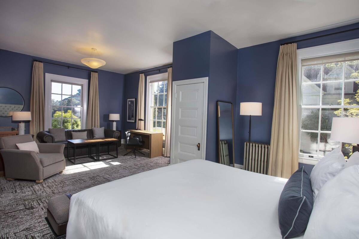 A hotel room with a bed, a desk, a chair, and a sofa. There is a window with curtains and a view of greenery outside.