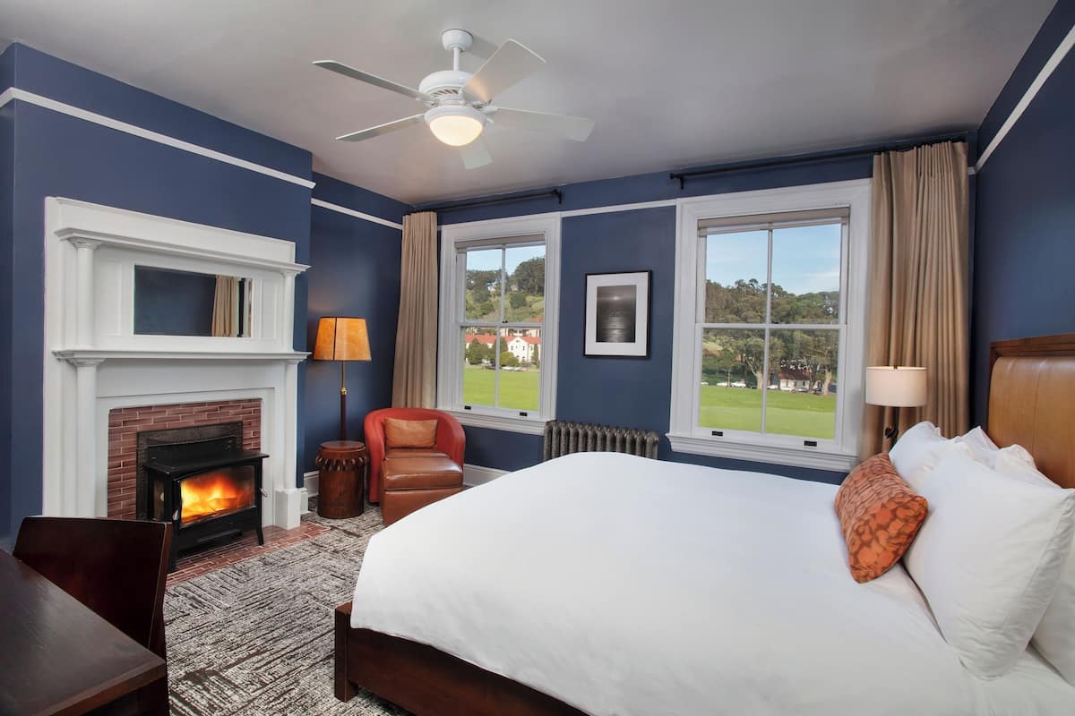 A bedroom with a fireplace, a bed, a desk, and a view of the outdoors.