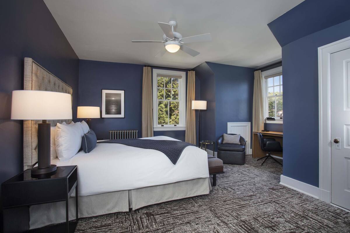 A bedroom with a bed, bedside lamps, a ceiling fan, a window with curtains, a desk, and a chair.