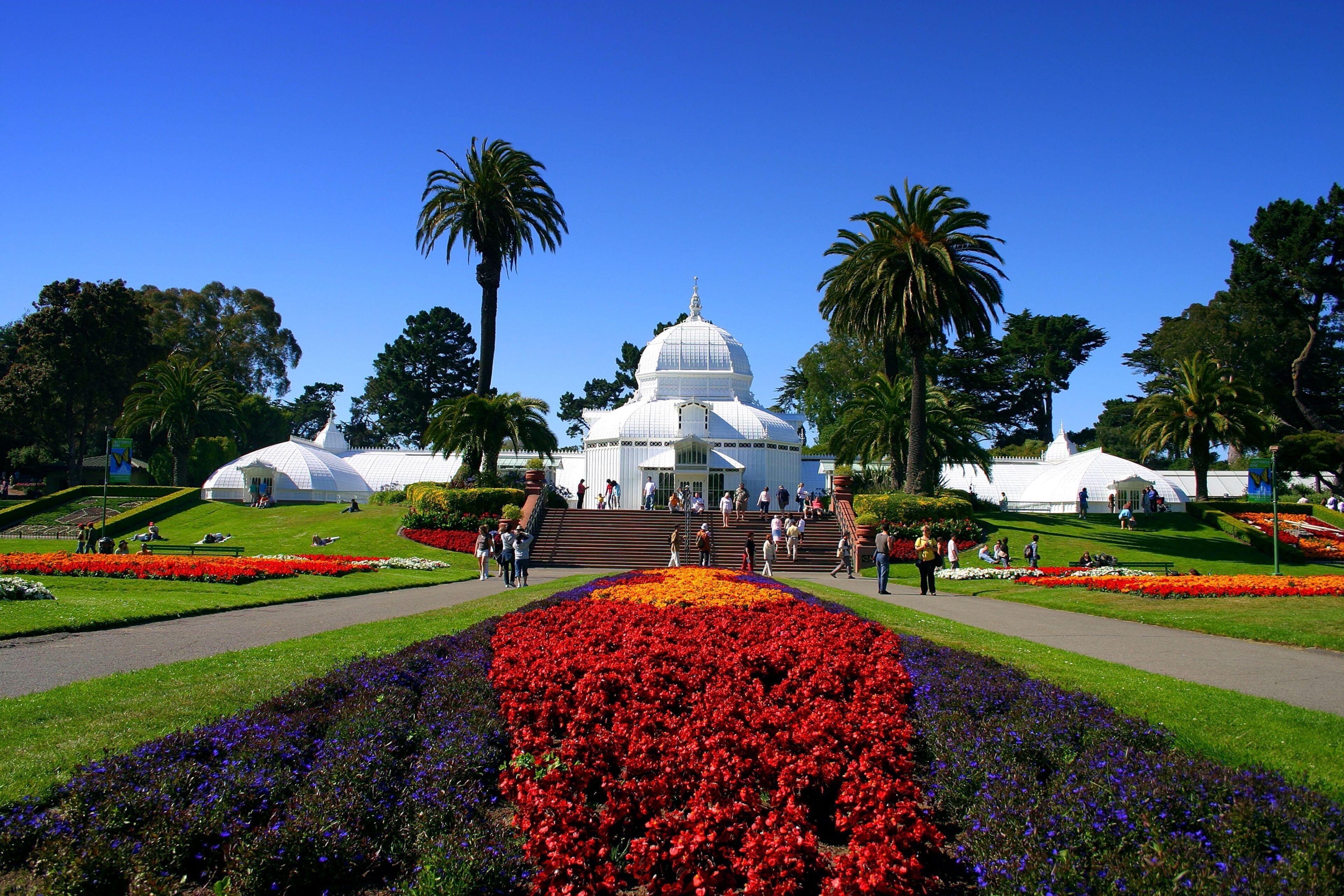 A large white dome building surrounded by a well-maintained garden with red and purple flowers, palm trees, and a clear blue sky.