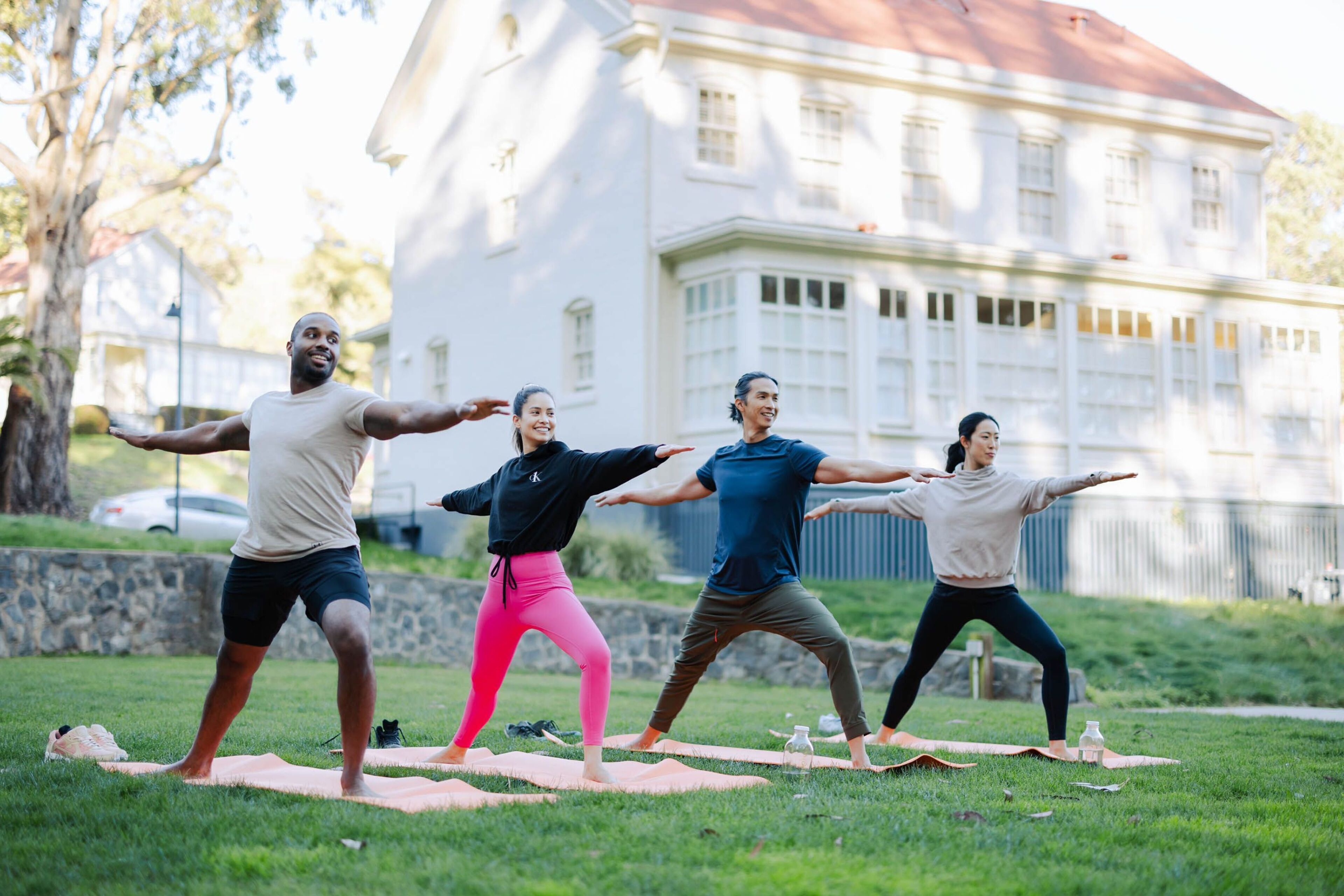 Quatro pessoas praticando yoga em tapetes, numa área gramada com um edifício ao fundo.