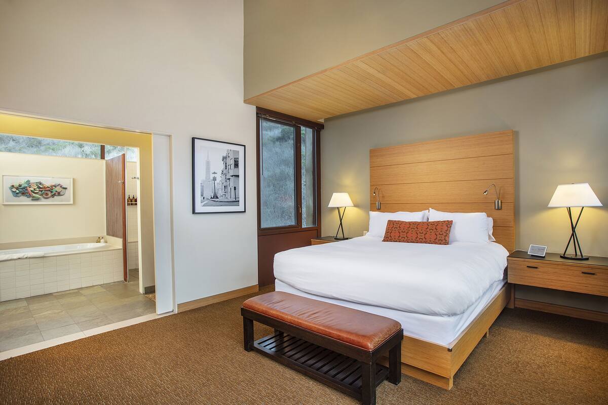 A modern hotel room with a large bed, a wooden headboard, two bedside lamps, a bench, and a view of the outside through a window.