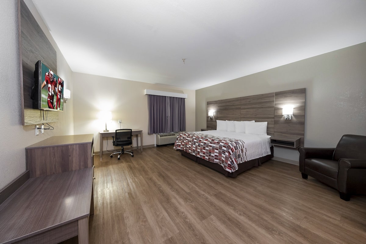 A hotel room with a wooden floor, a bed with a checkered bedspread, a desk with a chair, a TV mounted on the wall, and a nightstand.
