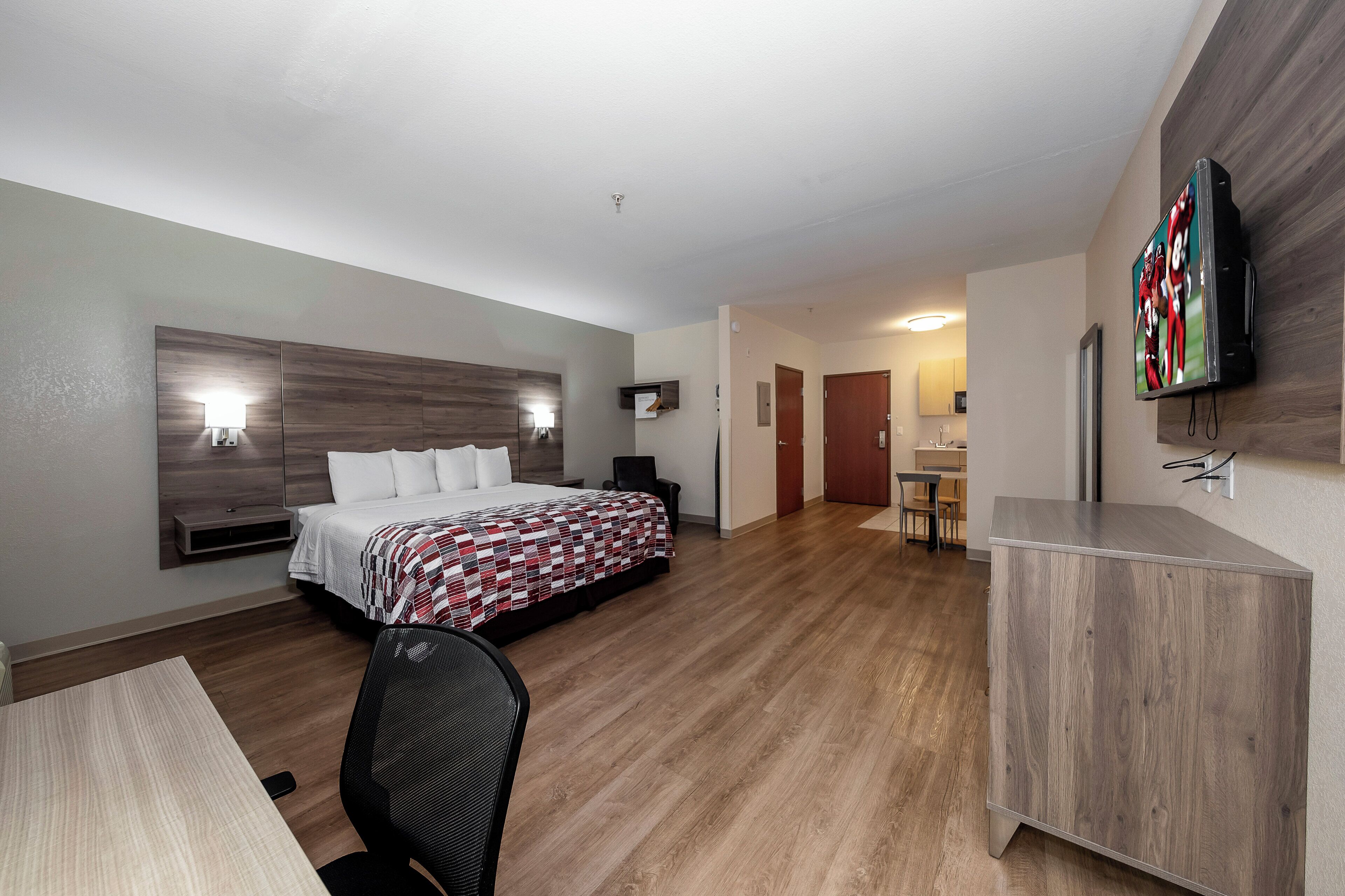 Quarto de hotel com cama, mesas de cabeceira, escrivaninha, cadeira, televisão na parede e banheiro com pia e espelho.