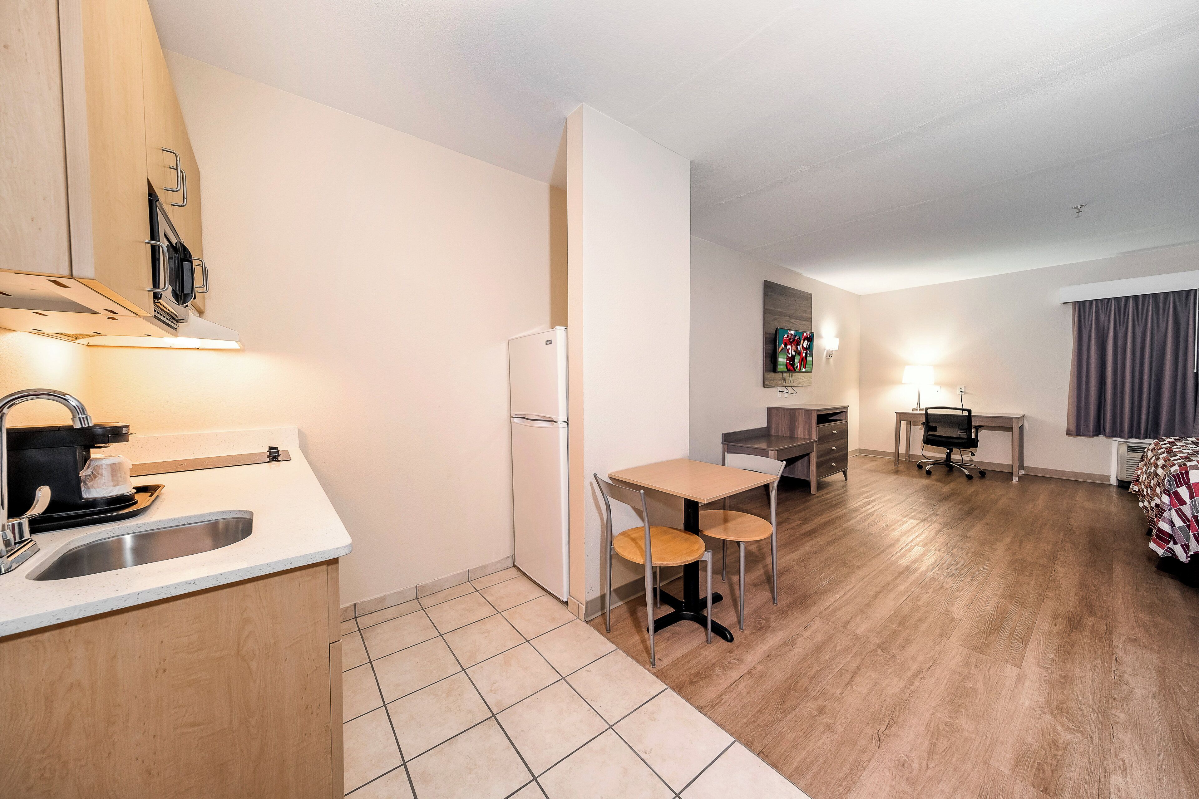 Cozinha compacta com pia, fogão e geladeira, pequena área de refeições com mesa e cadeiras, e quarto com cama e janela.