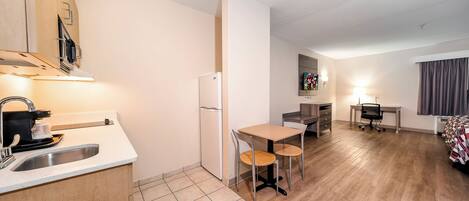 Cozinha compacta com pia, fogão e geladeira, pequena área de refeições com mesa e cadeiras, e quarto com cama e janela.