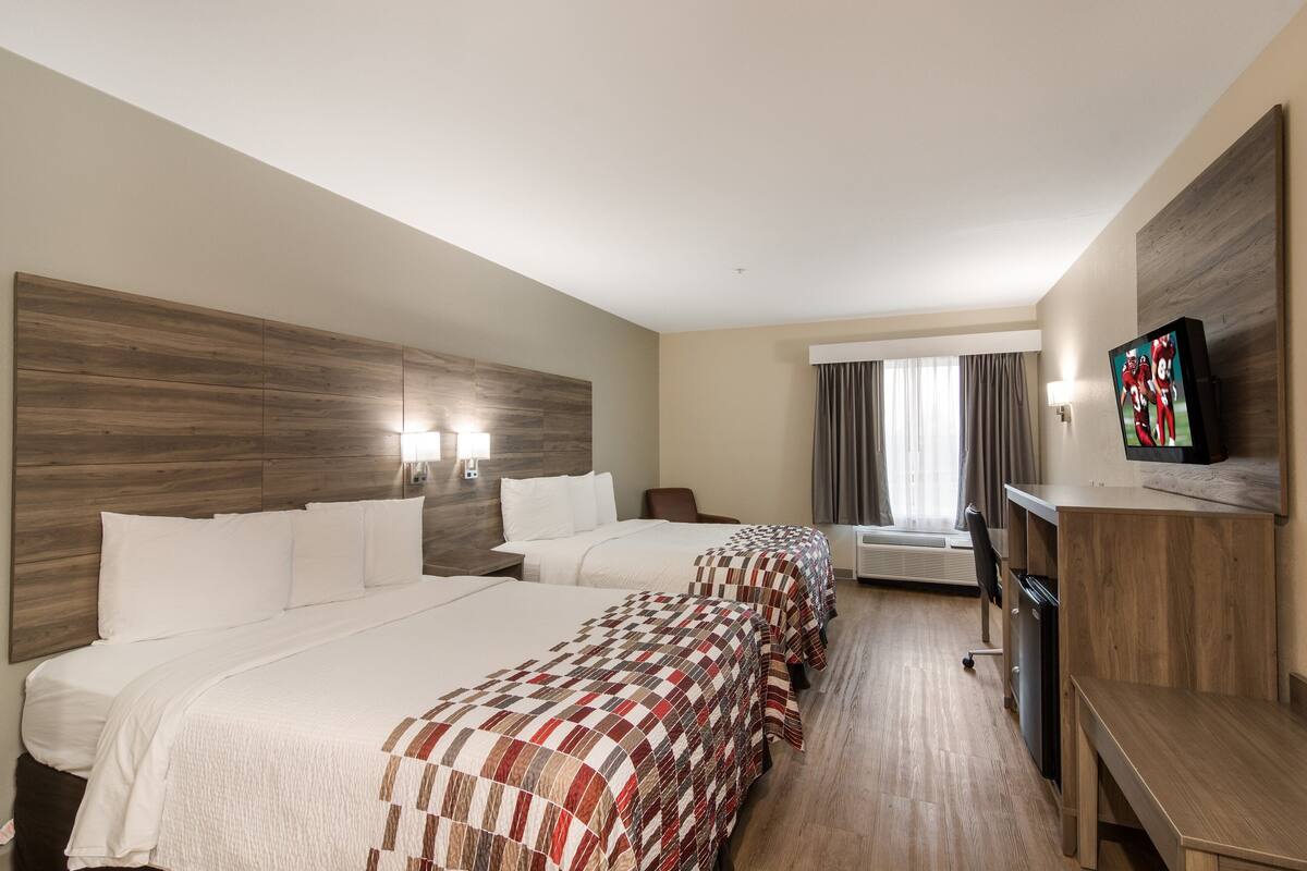 A hotel room with two beds, a desk, a TV, and a window with curtains.