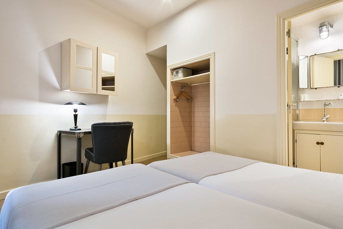 Quarto de hotel com duas camas, uma escrivaninha com cadeira, guarda-roupa e banheiro com pia e espelho.