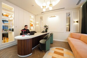 A reception area with a counter, a seated receptionist, a sofa, and a decorative vase with flowers. - Hotel Condal (Barcelona)