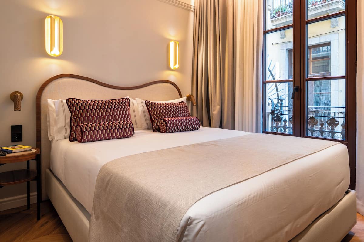 A neatly made bed with patterned pillows, a bedside table with a book, and a window with curtains.