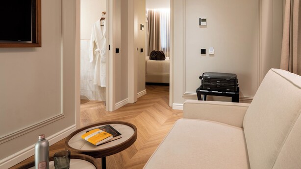 A hotel room with a sofa, a small round table with a book, a suitcase, and a robe hanging in the doorway. - Hotel Condal (Barcelona)
