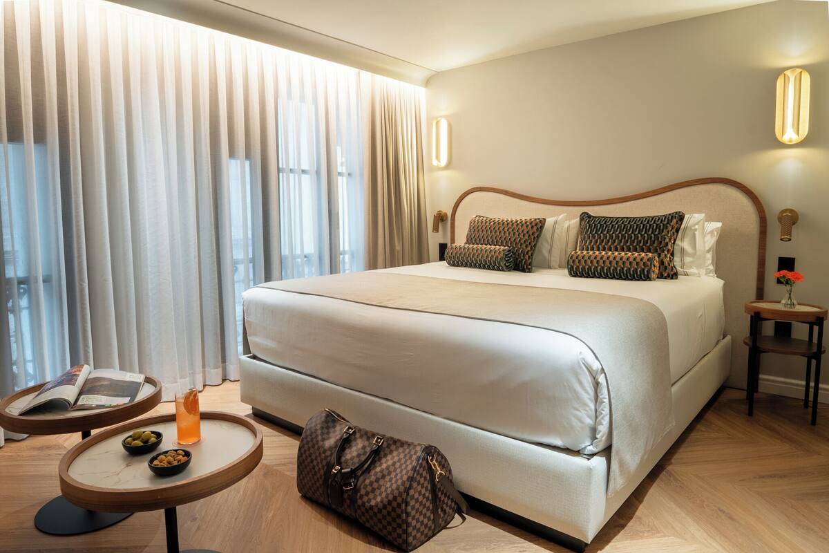 A hotel room with a large bed, a bedside table with a magazine and a drink, and a small table with snacks.