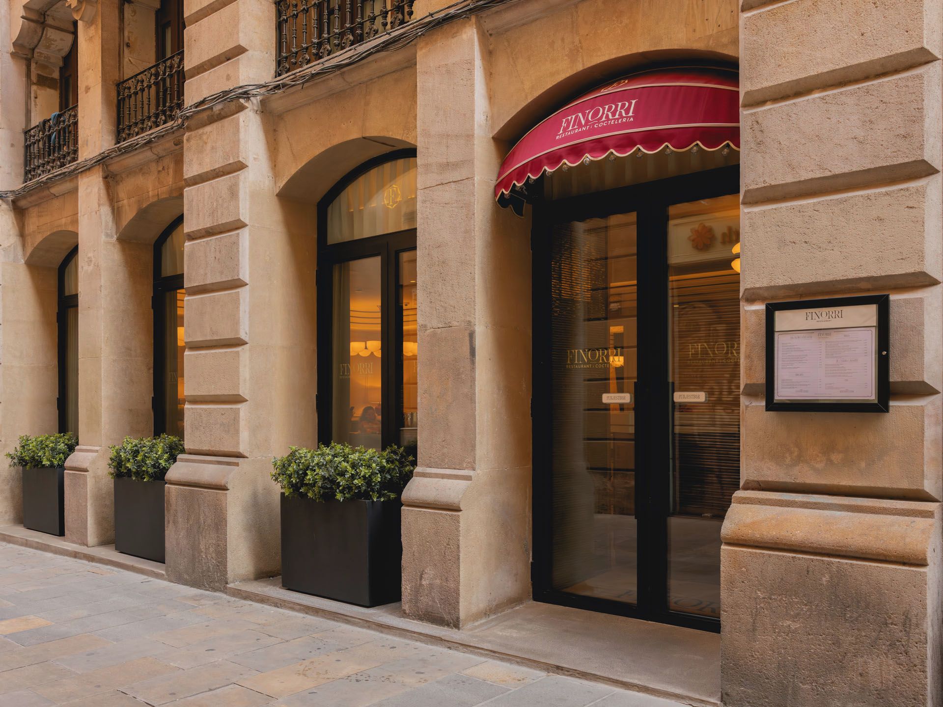 Fachada de un edificio con un restaurante llamado 'FINORRI', que presenta un toldo rojo, ventanas grandes y un menú enmarcado en la pared.