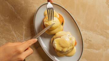 Une personne tient une fourchette et un couteau au-dessus d’une assiette contenant deux œufs pochés recouverts de sauce hollandaise.