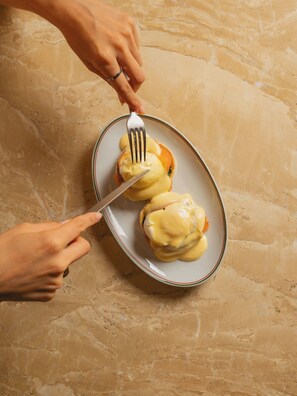 Une personne tient une fourchette et un couteau au-dessus d’une assiette contenant deux œufs pochés recouverts de sauce hollandaise.