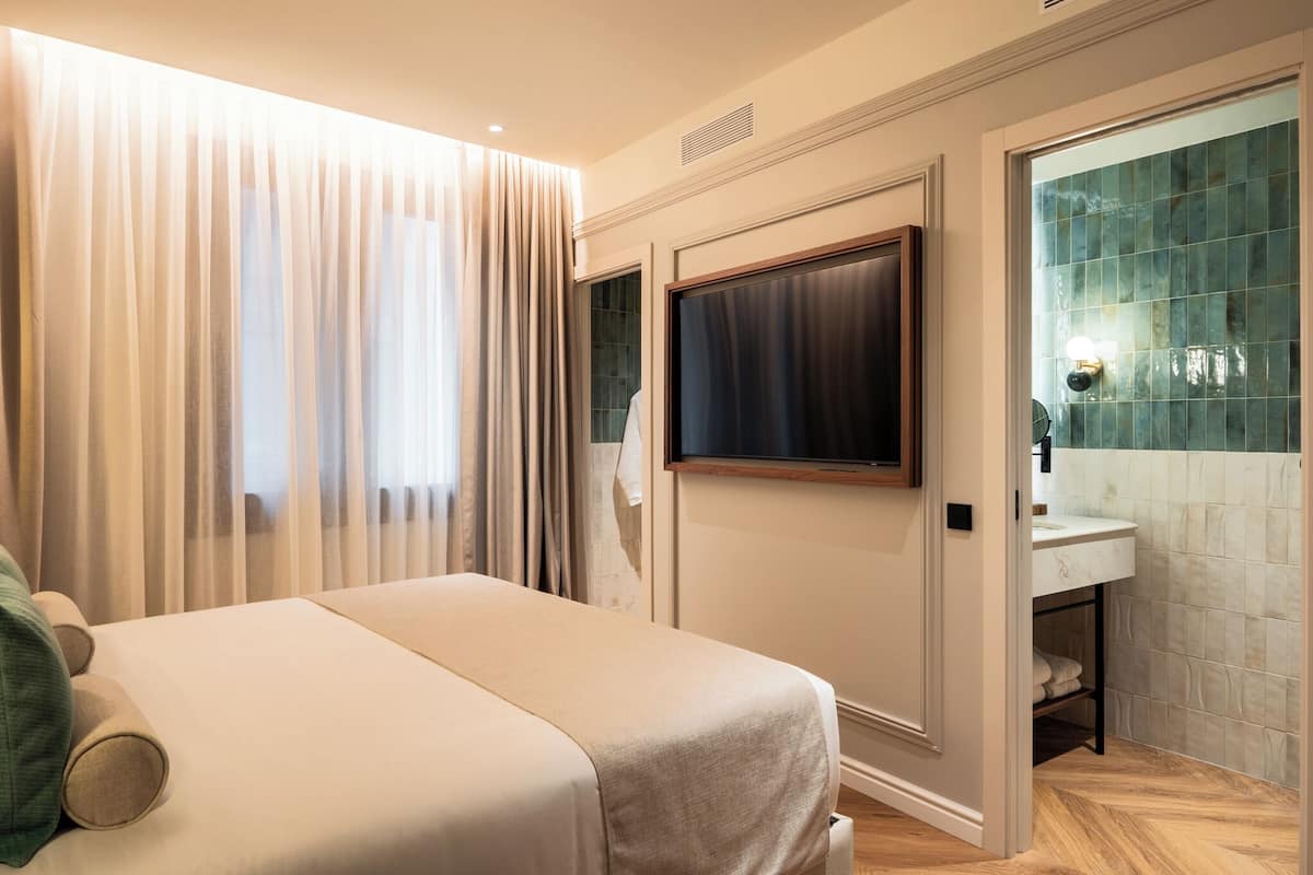 A hotel room with a bed, a TV mounted on the wall, a bathroom with a tiled wall, and a door leading to another room.