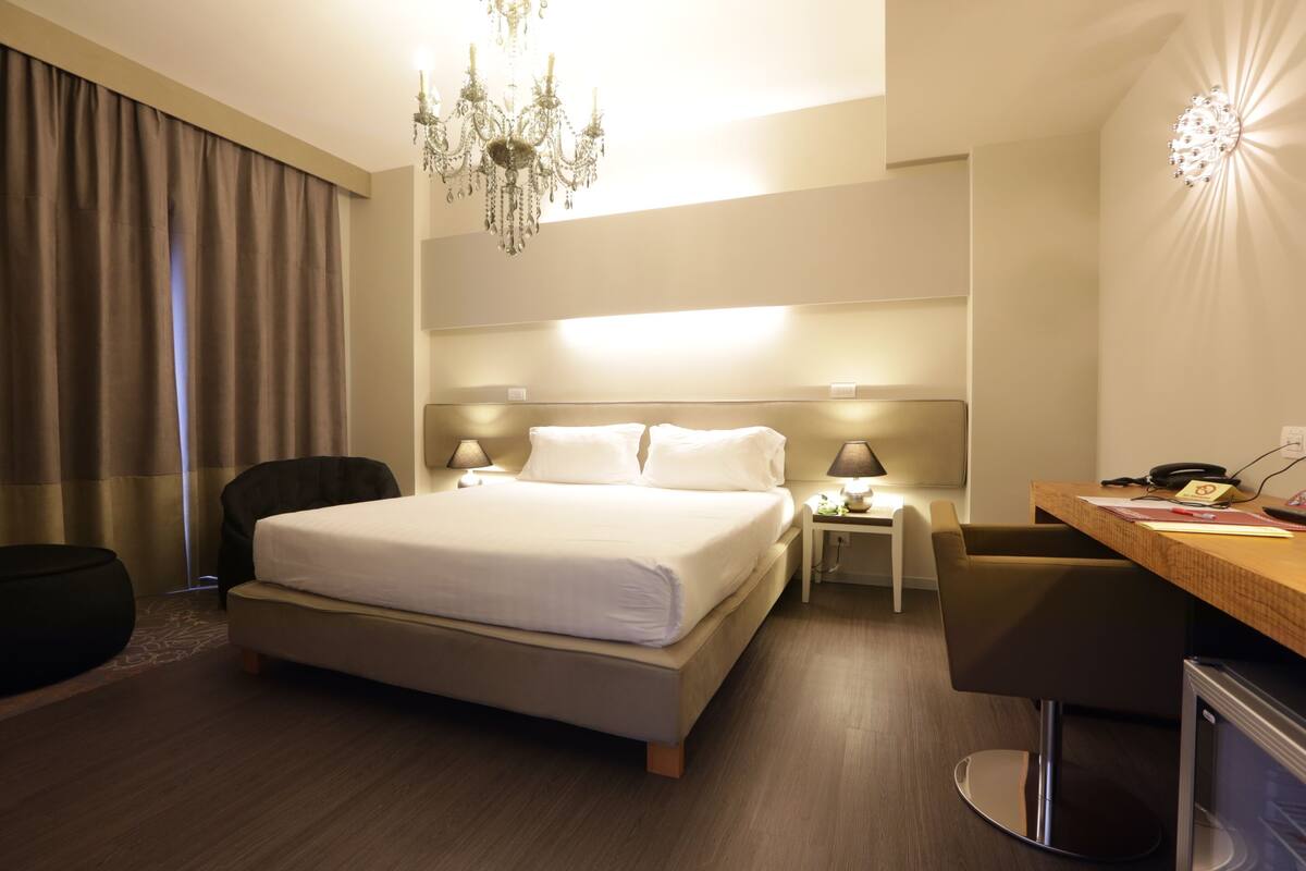 Une chambre d’hôtel avec un grand lit, un bureau, une chaise, une lampe de bureau, une table de chevet, un lustre et un téléviseur.