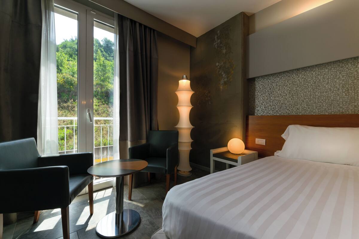 Une chambre d’hôtel avec un lit, un bureau, une chaise, une lampe et une vue sur la verdure par la fenêtre.