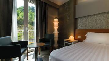 Une chambre d’hôtel avec un lit, un bureau, une chaise, une lampe et une vue sur la verdure par la fenêtre.
