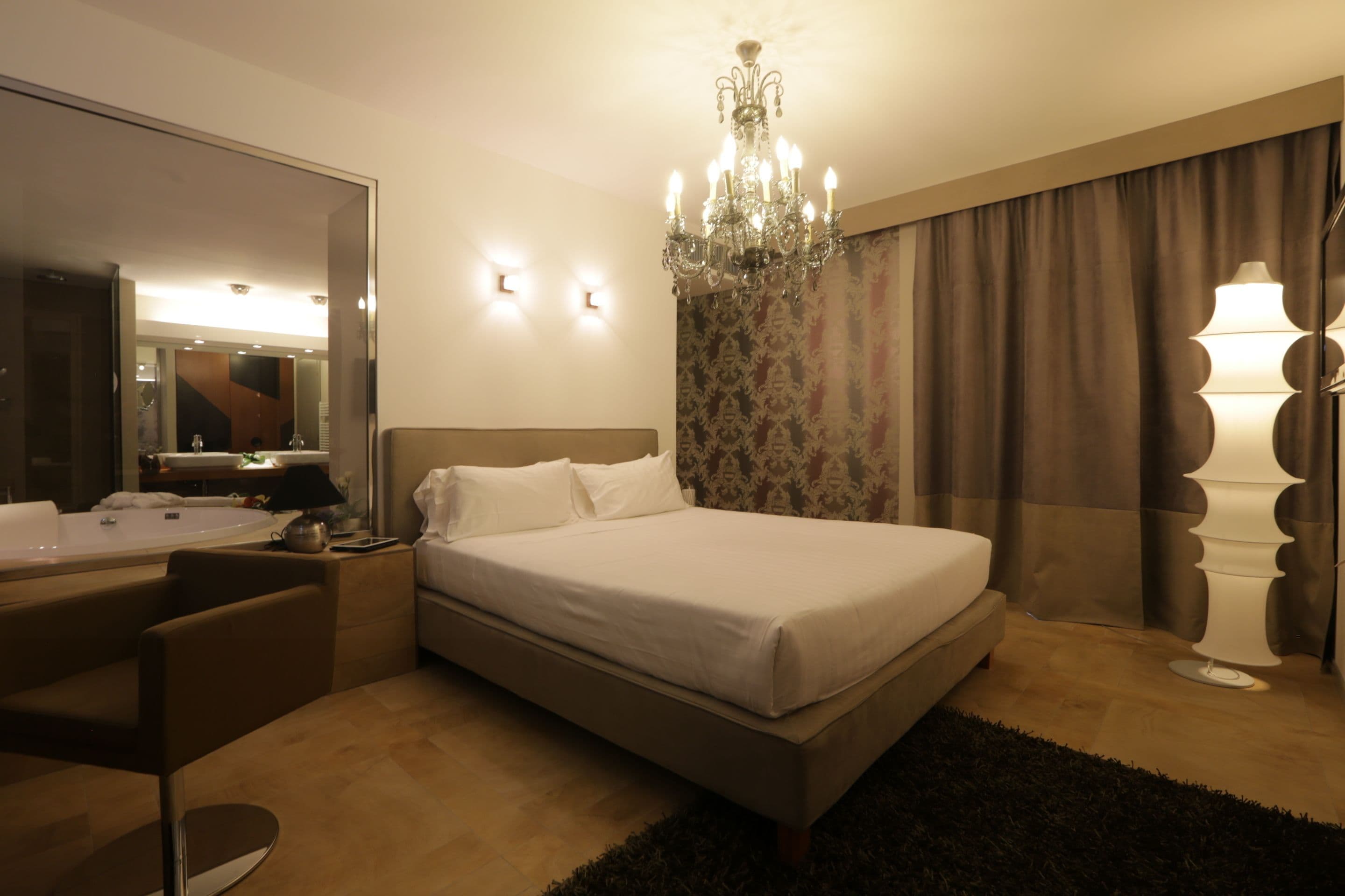 A modern hotel room with a large bed, a sitting area with a chair, a vanity with a mirror, a chandelier, and a large window with curtains.