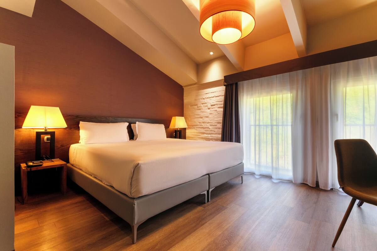 Une chambre d’hôtel moderne avec un grand lit, deux lampes de chevet, une table de chevet en bois, une chaise noire et une vue sur l’extérieur grâce à de grandes fenêtres.