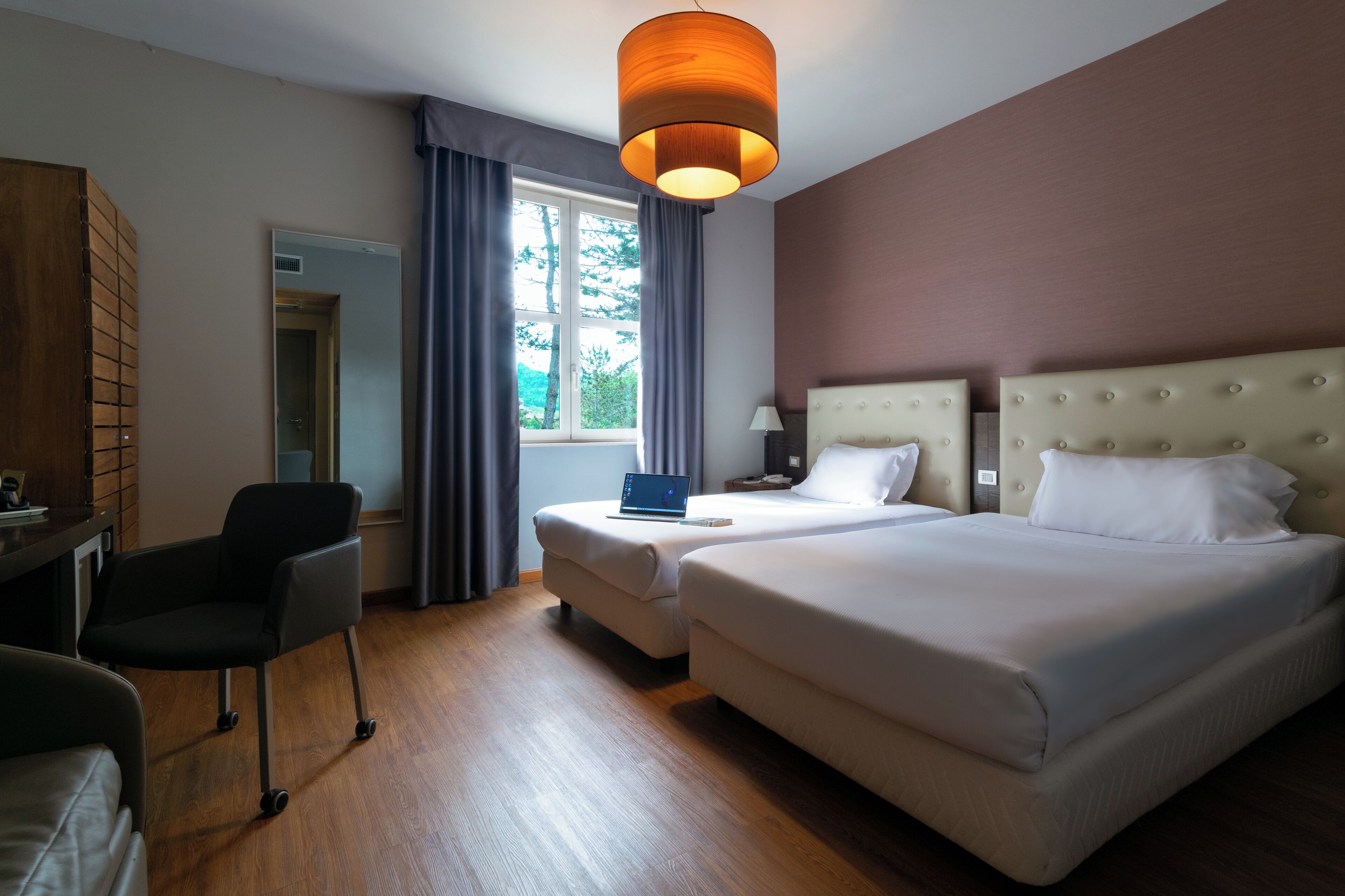 Habitación de hotel con una cama grande, un escritorio con silla, una computadora portátil y una ventana con cortinas.