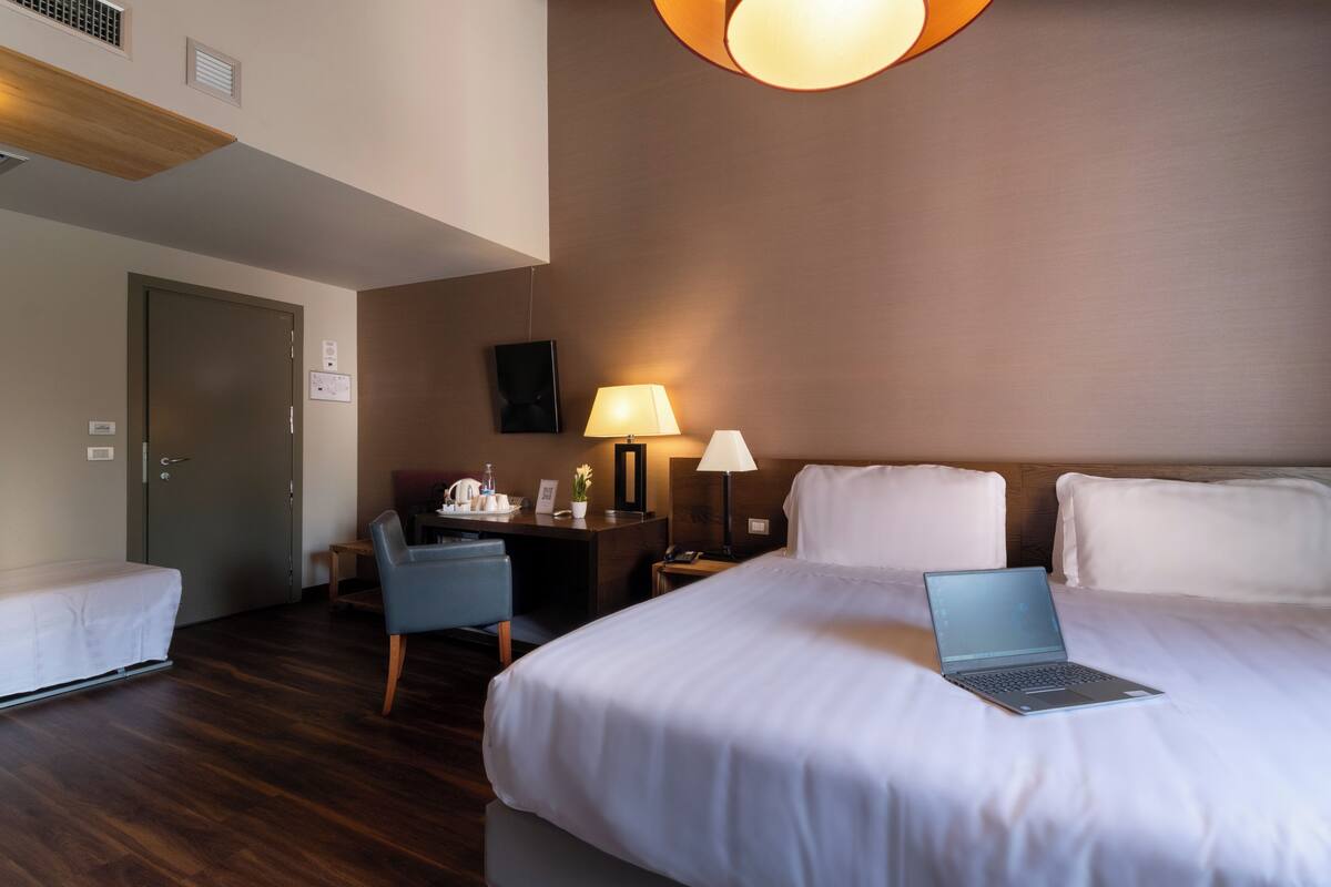 Une chambre d’hôtel avec un lit, un bureau avec un ordinateur portable, une chaise, une lampe et une porte.