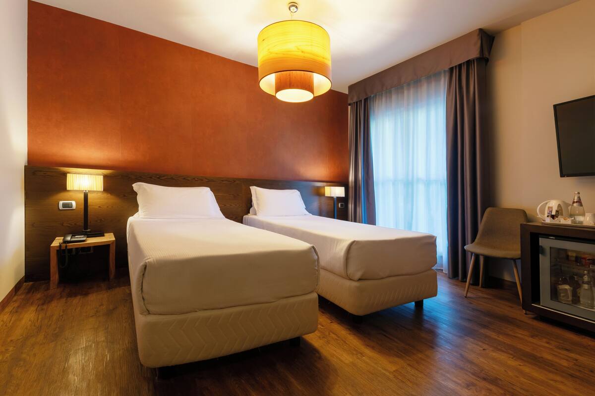 Une chambre d’hôtel avec deux lits simples, une tête de lit en bois, une table de chevet avec une lampe, un bureau avec une chaise et un téléviseur fixé au mur.