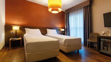 Camera d'albergo con due letti singoli, una testiera in legno, un comodino con una lampada, una scrivania con una sedia e un televisore a parete.