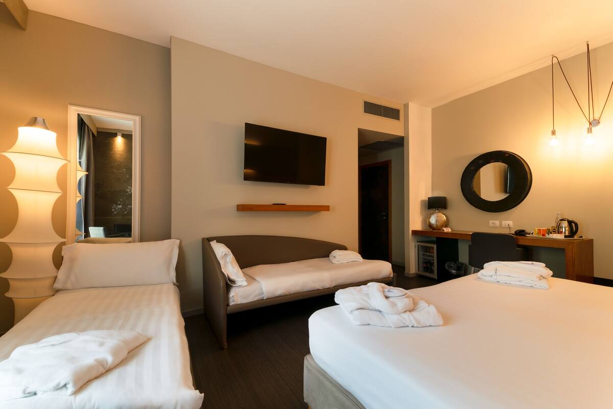 Une chambre d’hôtel avec deux lits simples, un grand lit, une télévision, un bureau avec un miroir et une lampe.