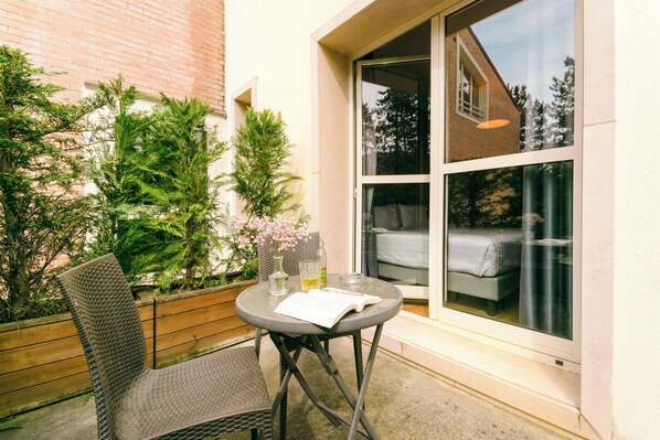A small patio with a round table, two chairs, a potted plant, and a window with a view of a bed inside.