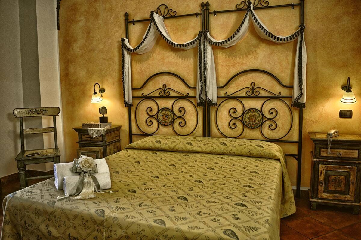 Una cama bien hecha con una colcha estampada, un cabecero decorativo con cortinas, mesitas de noche con lámparas y una pequeña silla de madera.