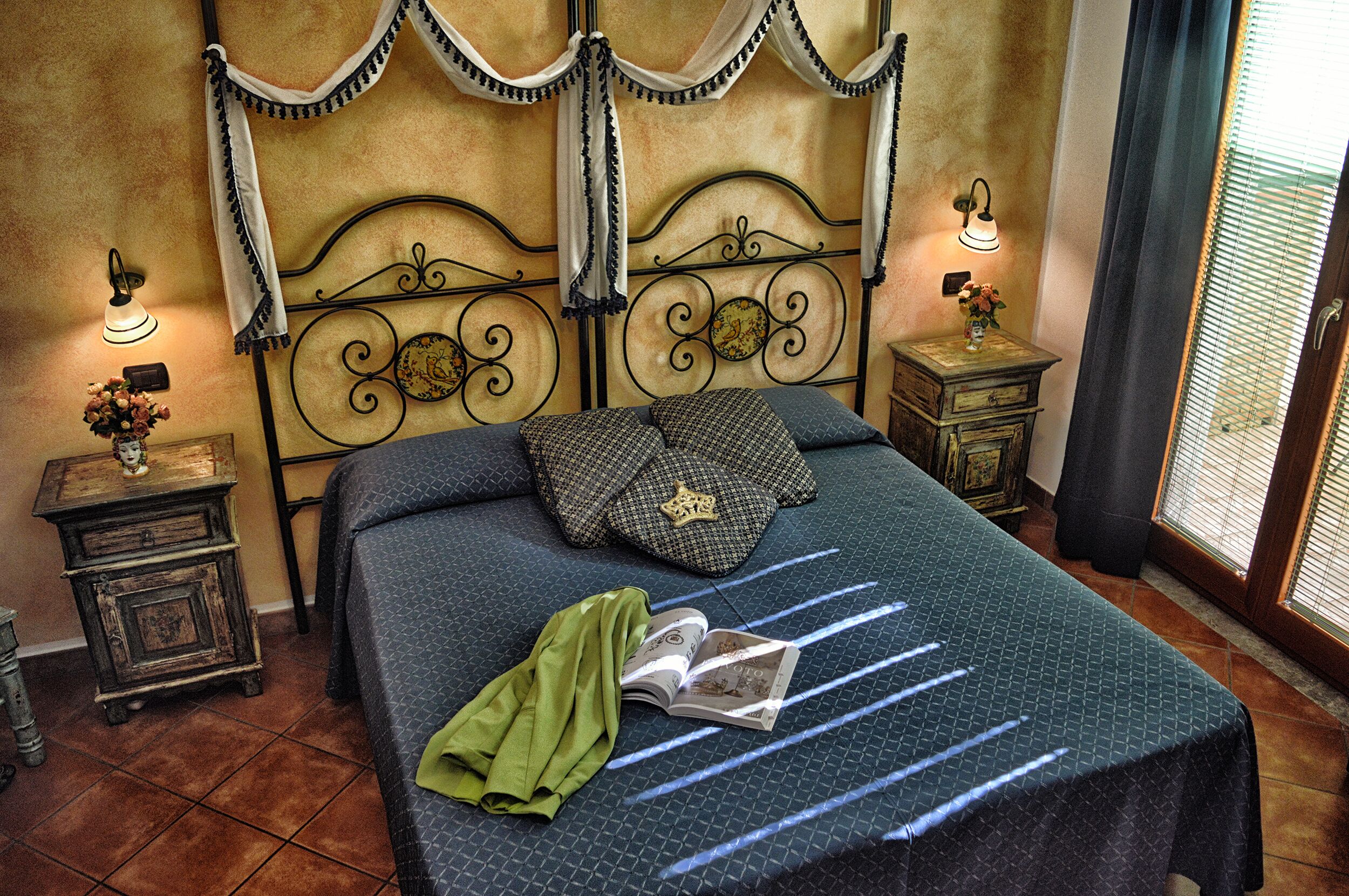 Un lit bien fait, recouvert d’une couvre-lit à motifs, avec un tissu vert et un livre. Il y a deux tables de chevet, chacune avec une lampe, et l’une d’elles est ornée d’un objet décoratif.