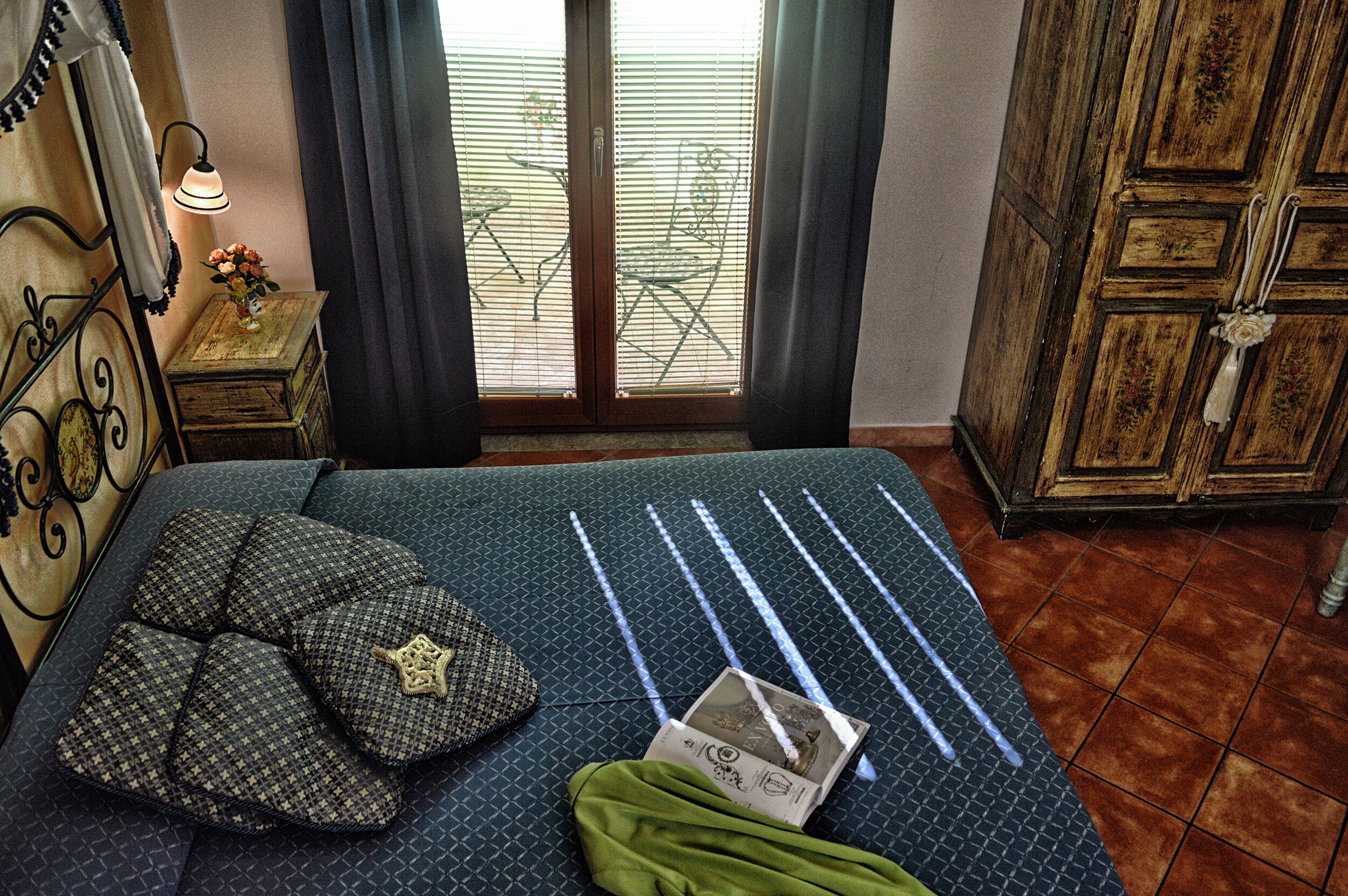 Ein Bett mit gemusterten Kissen, ein Nachttisch mit einer Lampe und Blick durch ein Fenster mit Jalousien.