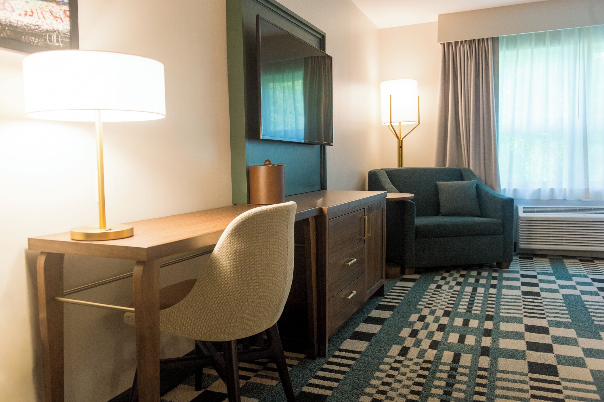 A hotel room with a wooden desk, a chair, a sofa, a large mirror, and a patterned rug.