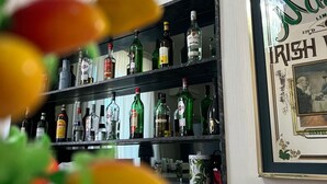 A bar area with shelves of liquor bottles, a framed sign reading "Nab Irish," and a blurred foreground of green and yellow objects. - 38 Aira Hotels (Palermo)
