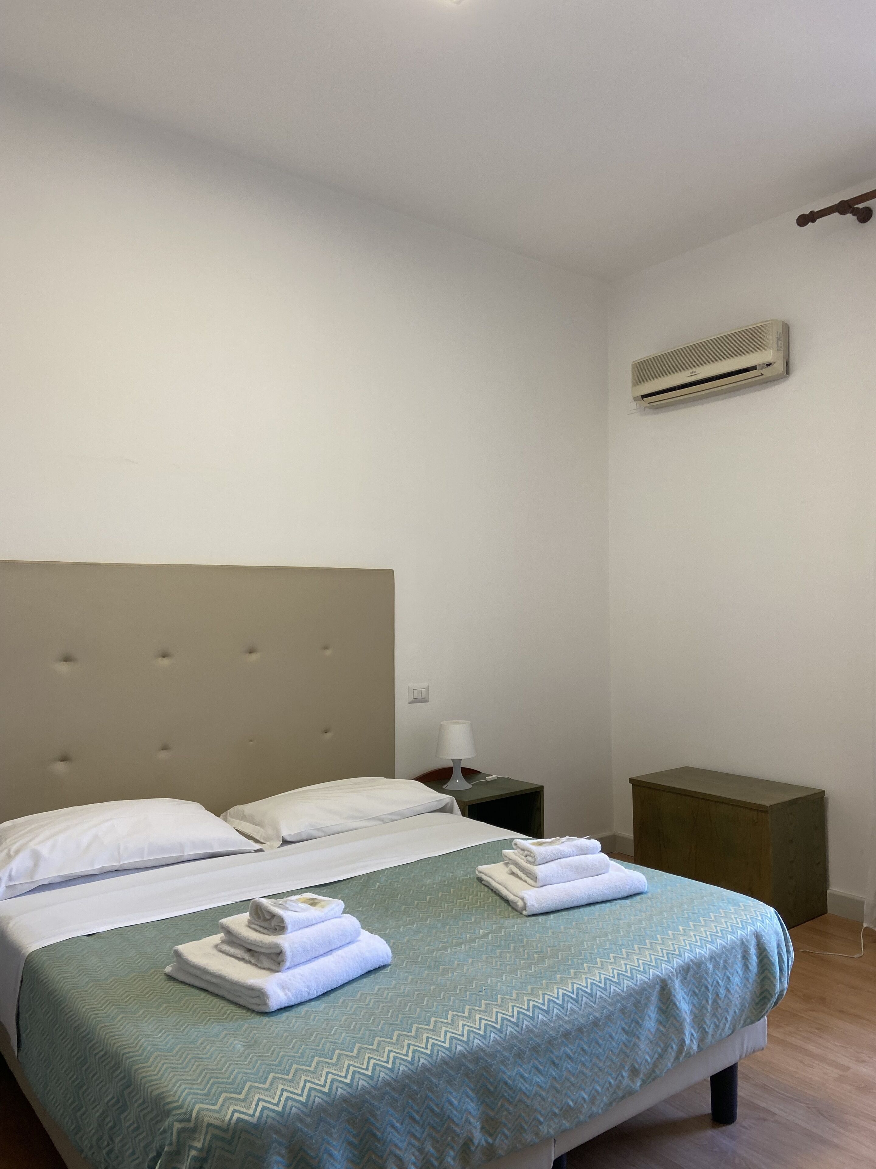 Habitación de hotel con cama doble, mesita de noche con lámpara, unidad de aire acondicionado y un pequeño armario de madera.