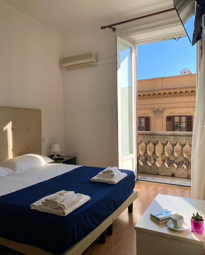 A hotel room with a bed, a nightstand, a table with a cup and a book, and a balcony with a railing.