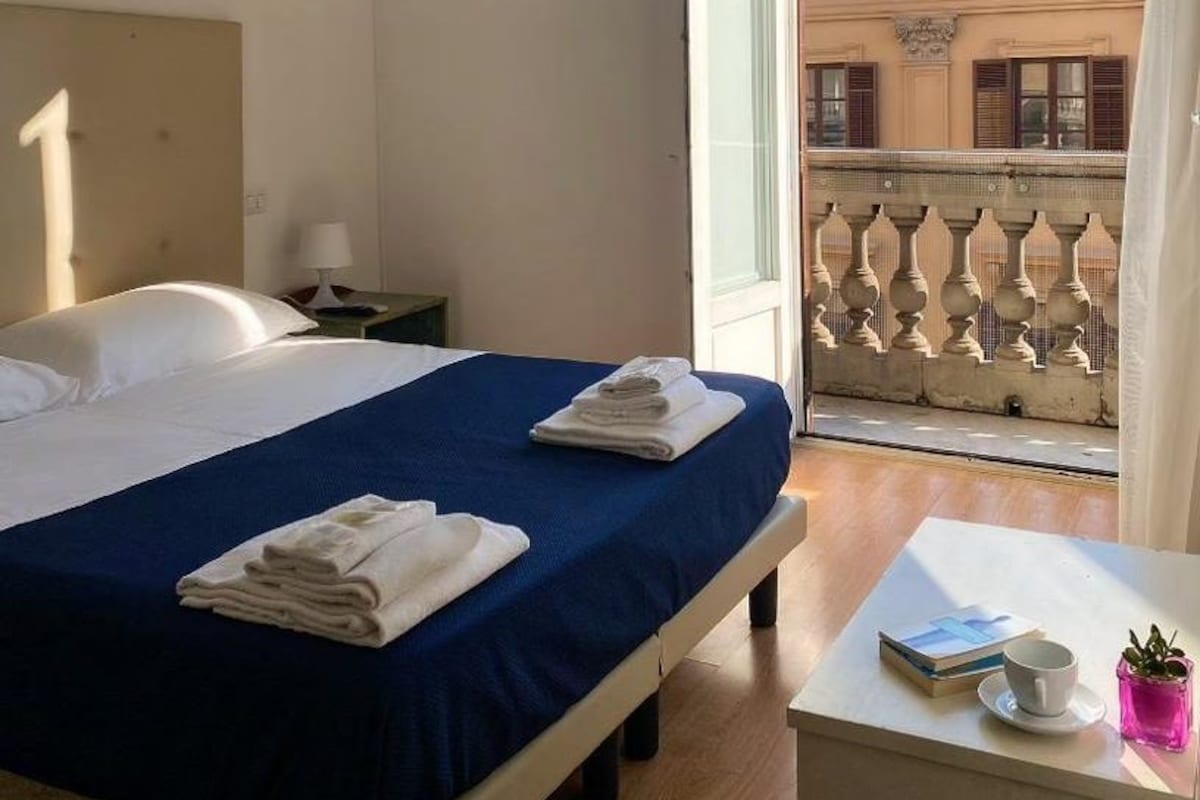 Una camera d'albergo con un letto, un comodino, un tavolo con una tazza e un libro, e un balcone con ringhiera.