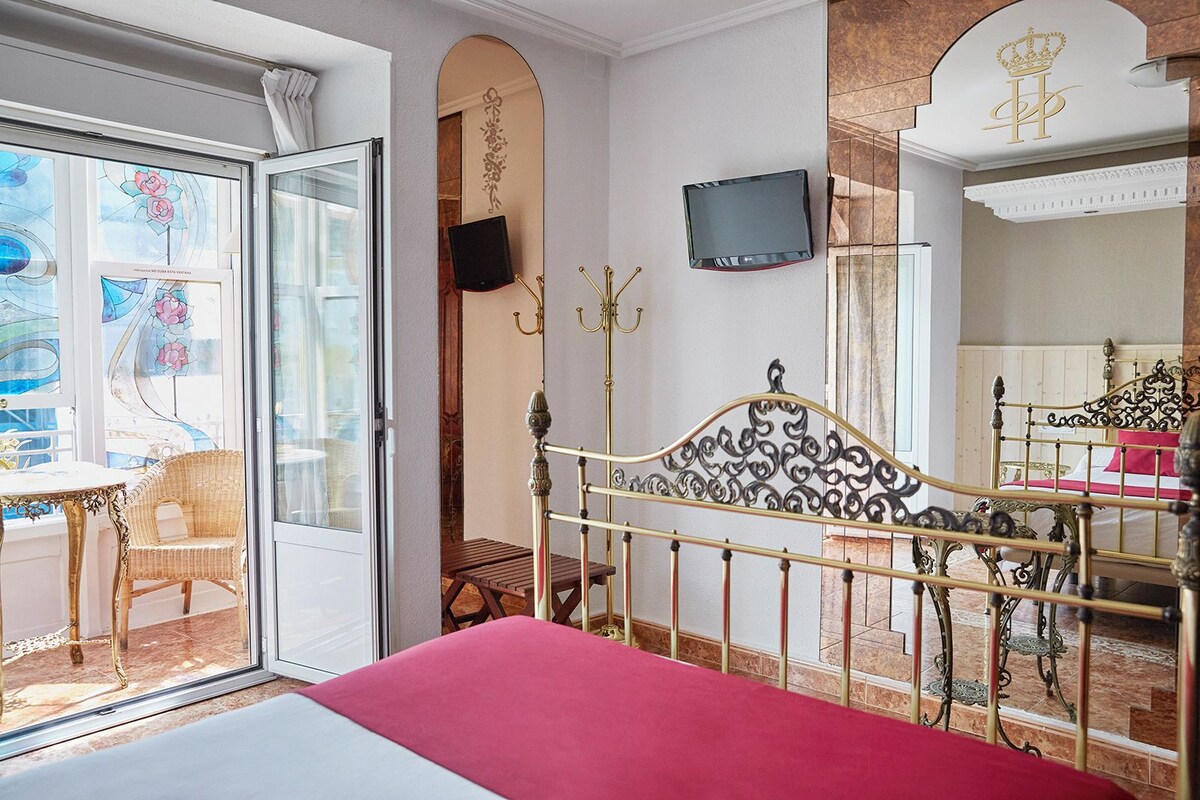 A hotel room with a bed, a TV, a balcony with a chair, and a decorative mirror.