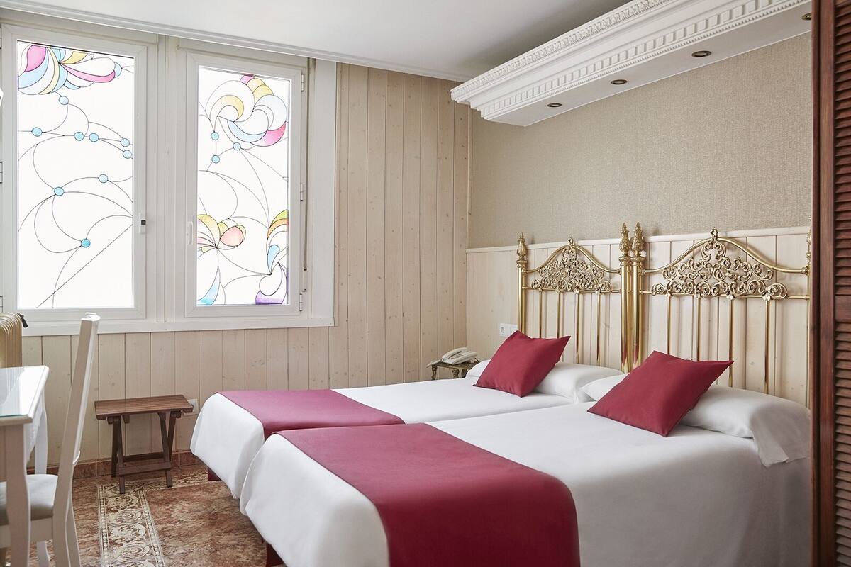 A hotel room with two beds, a desk, and a window with stained glass.