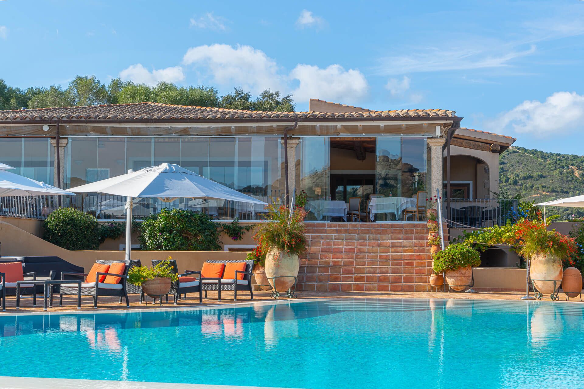 A poolside area with lounge chairs and umbrellas, a brick wall, and a building with large windows and a balcony.