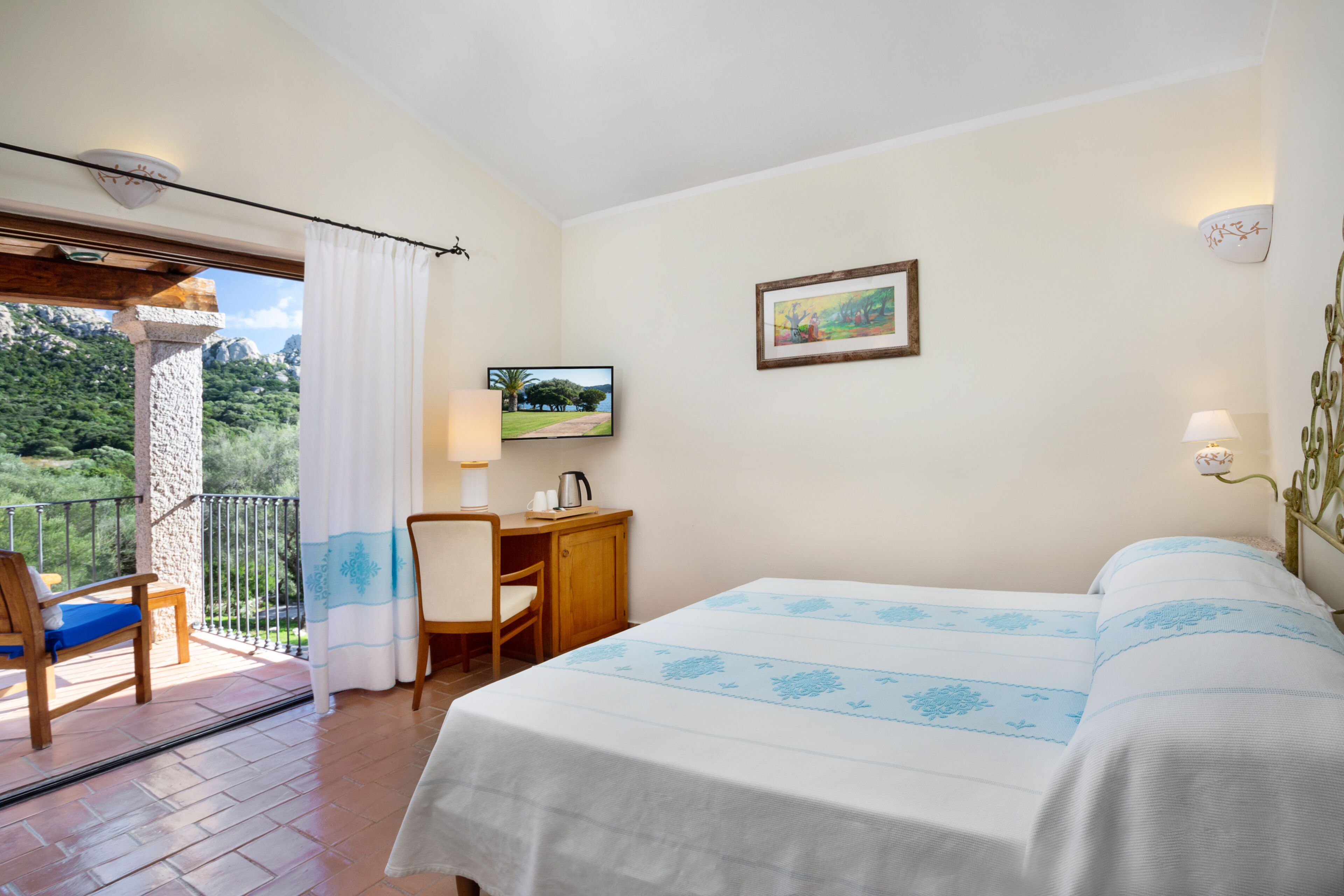 A bedroom with a bed, a desk with a chair, a TV, and a balcony with a view of greenery.