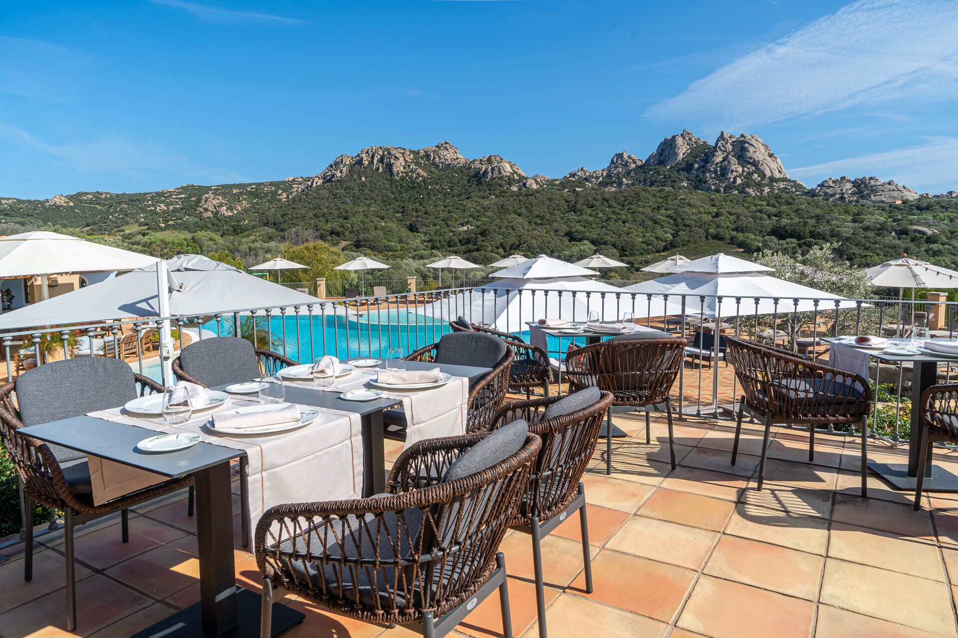 An outdoor dining area with tables and wicker chairs, overlooking a pool and mountains.