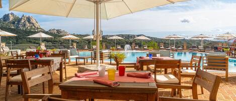 A poolside area with wooden tables and chairs, umbrellas, and a view of a mountainous landscape.