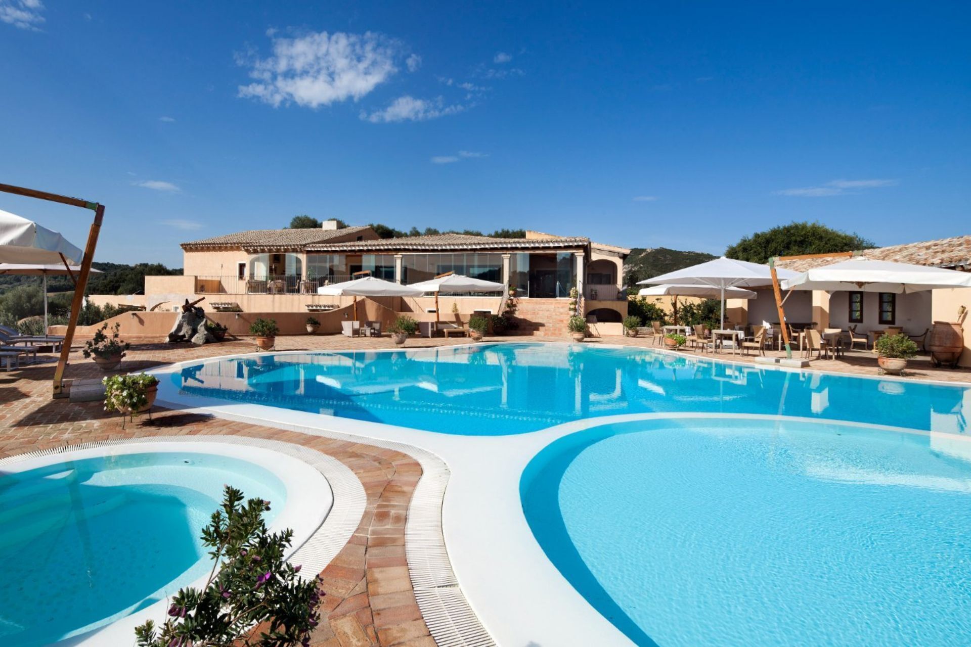 A swimming pool with a curved design, surrounded by lounge chairs and umbrellas, and a building with a tiled roof in the background.