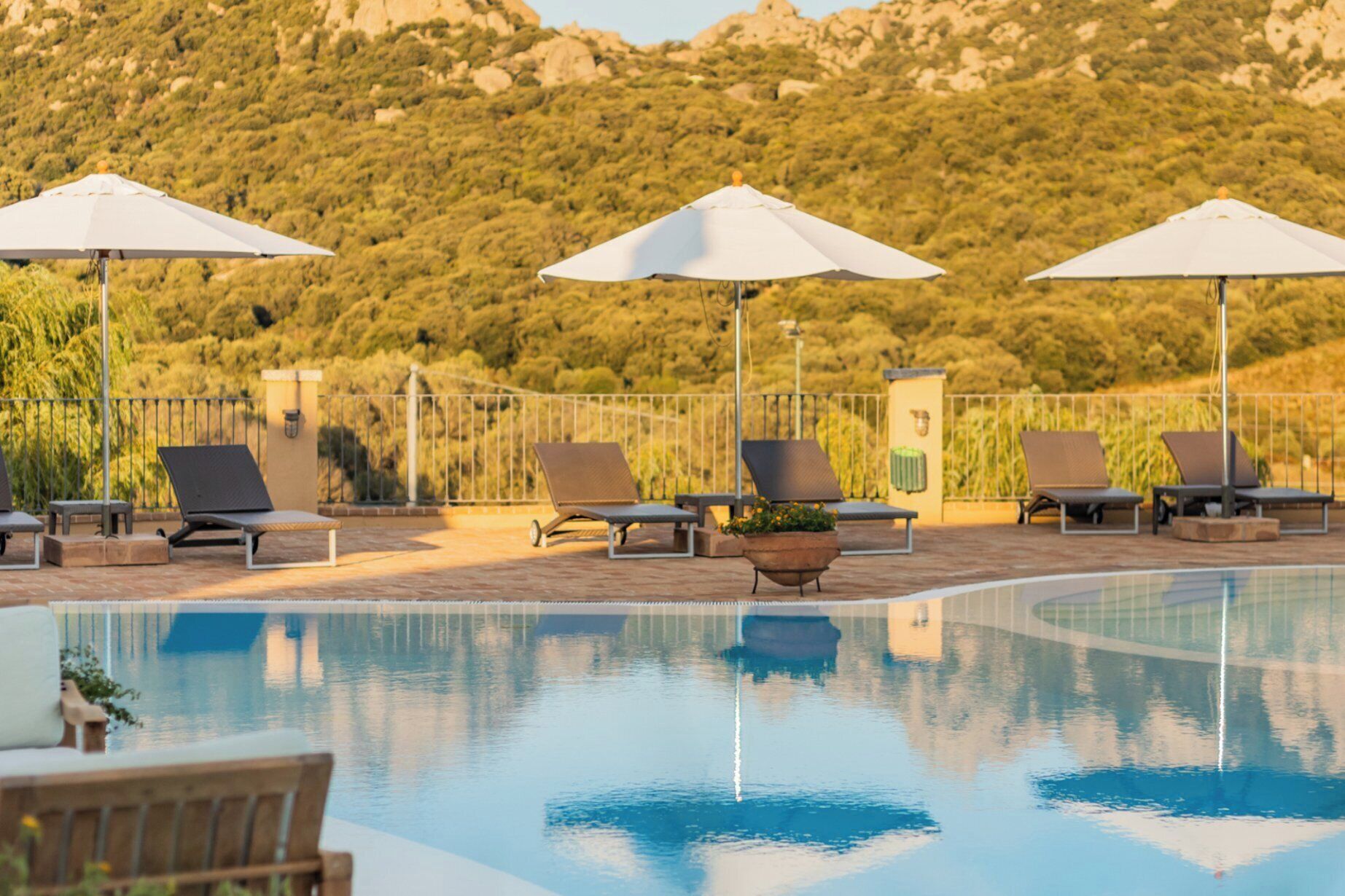 A pool area with lounge chairs and umbrellas, surrounded by a fence and a mountainous landscape.