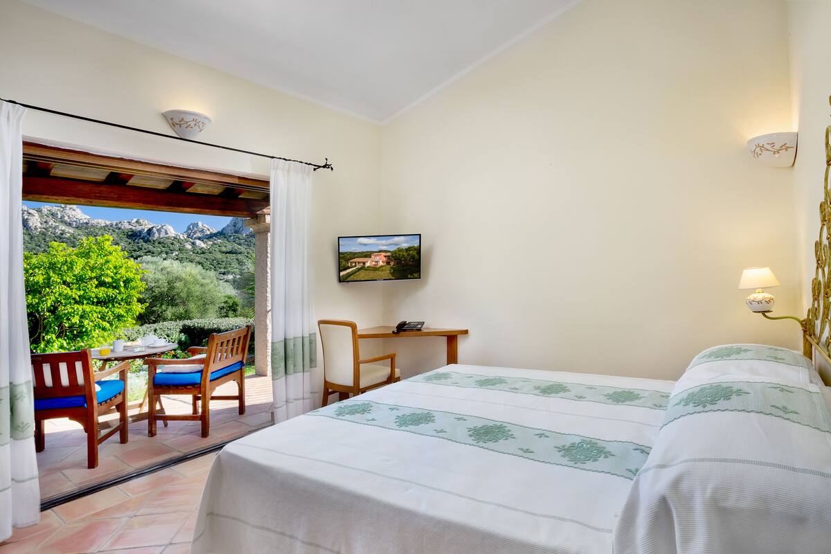 Ein Schlafzimmer mit einem Bett, einem Schreibtisch und einem Stuhl. Ein Fenster bietet Blick auf Berge und Bäume.