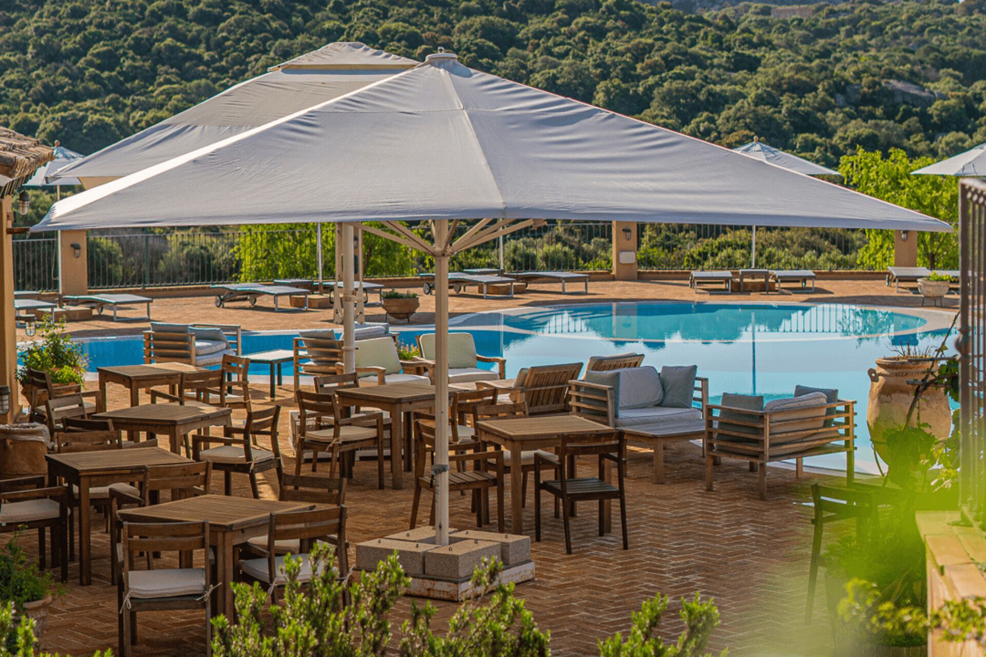 Zona de piscina con mesas y sillas de madera, sombrillas y vista a una alberca.